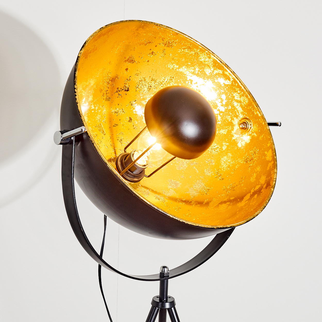 Eine schwarze Stehlampe mit großem, goldenem Innenschirm. Eine leuchtende Glühbirne ist darin. Der Sockel ist ein Stativ. Der Hintergrund ist weiß.