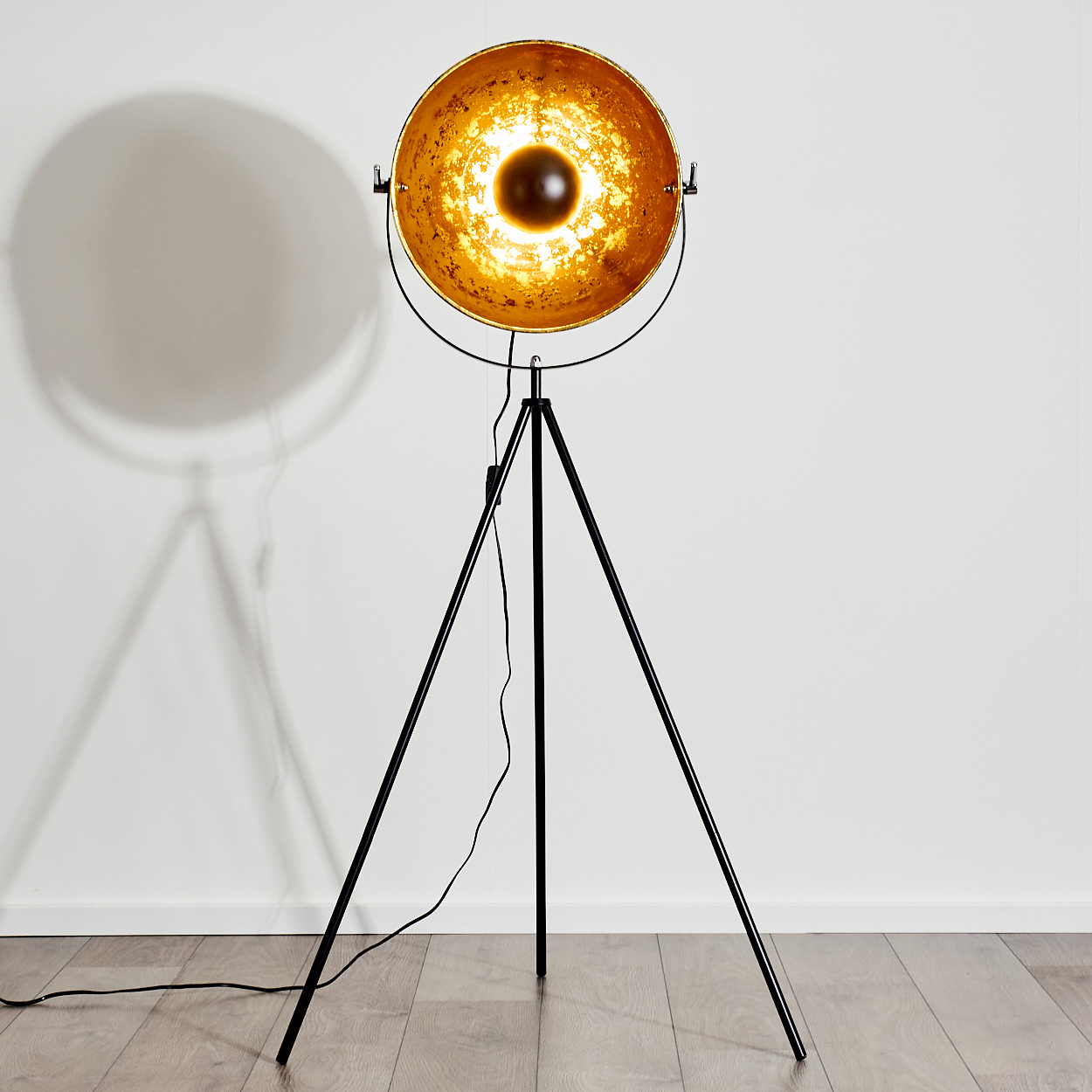 Eine schwarze Stativ-Stehlampe mit einem großen, goldfarbenen Lampenschirm. Sie steht vor einer weißen Wand und wirft einen Schatten.
