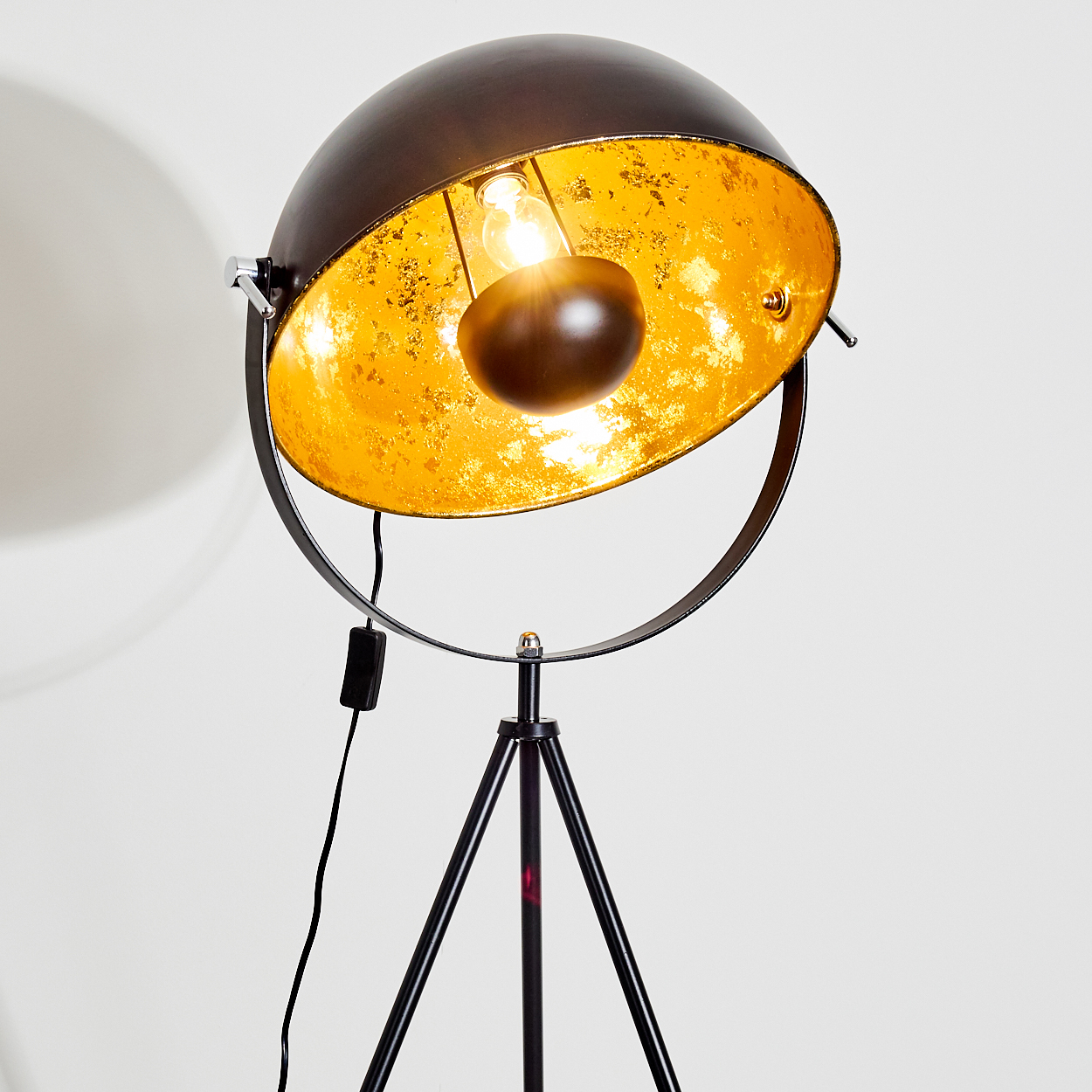 Eine schwarz-goldene Stehlampe. Der Lampenschirm ist eine dunkle Kuppel mit goldener Innenseite. Sie steht auf einem schwarzen Dreibein-Sockel.