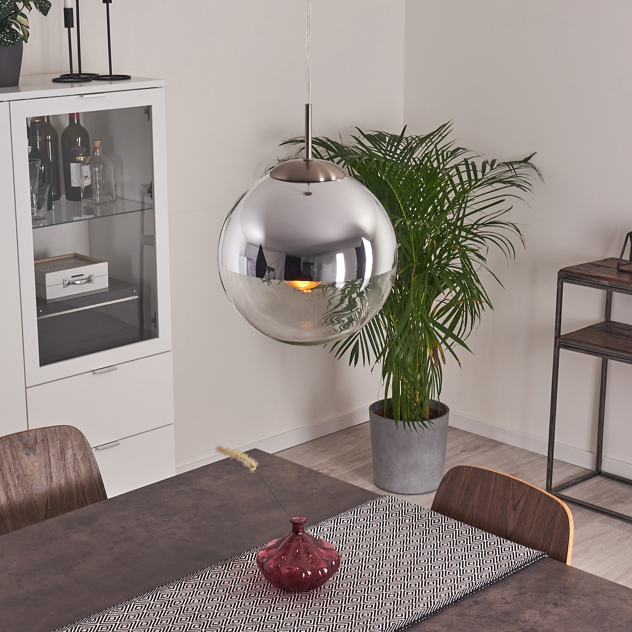 Ein modernes Esszimmer mit einer kugelförmigen Pendelleuchte, einem Tisch, Stühlen, einer Pflanze und einem Schrank mit Flaschen.