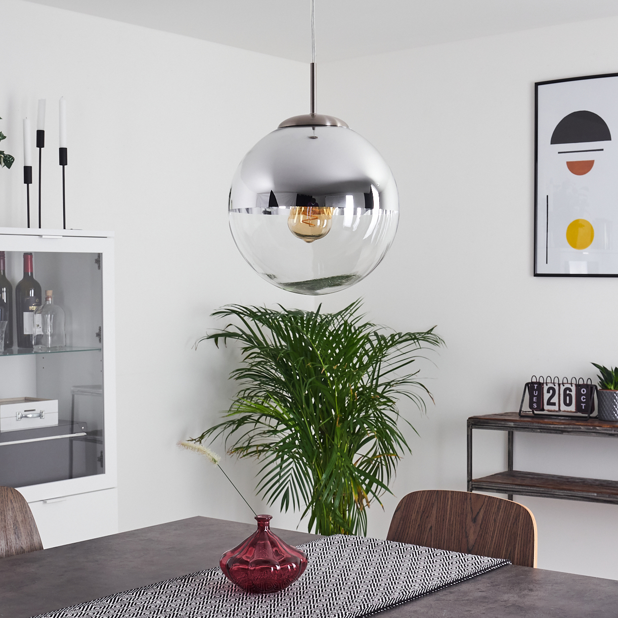 Ein modernes Esszimmer mit einer reflektierenden Pendelleuchte, einem Tisch und einer Pflanze. Auf dem Tisch steht eine rote Vase.