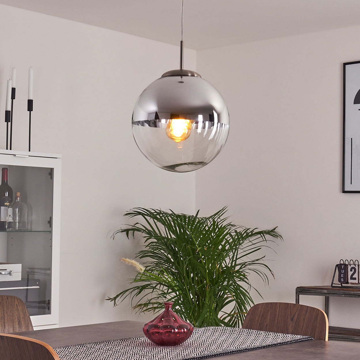 Ein modernes Esszimmer mit einer kugelförmigen Pendelleuchte, Pflanzen, einer roten Vase und einem gedeckten Tisch.