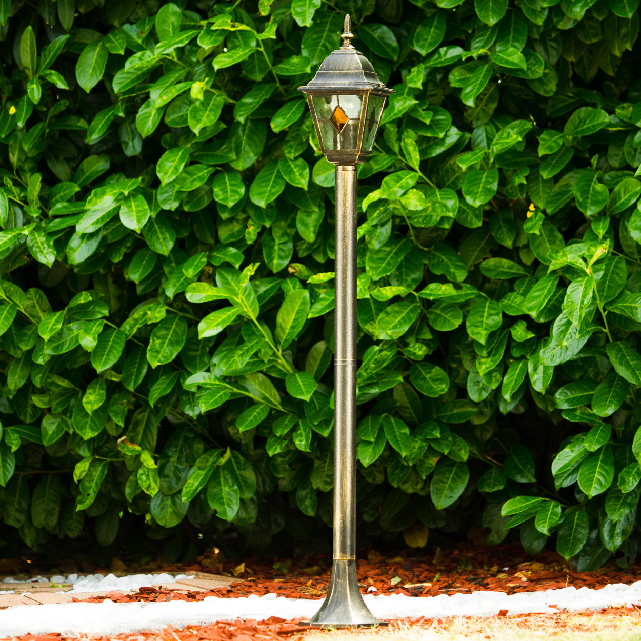 Eine dekorative Außenlampe steht vor einer dichten, grünen Hecke. Die Lampe ist bronzefarben und ihr Licht ist an. Weiße Steine und Mulch am Sockel.