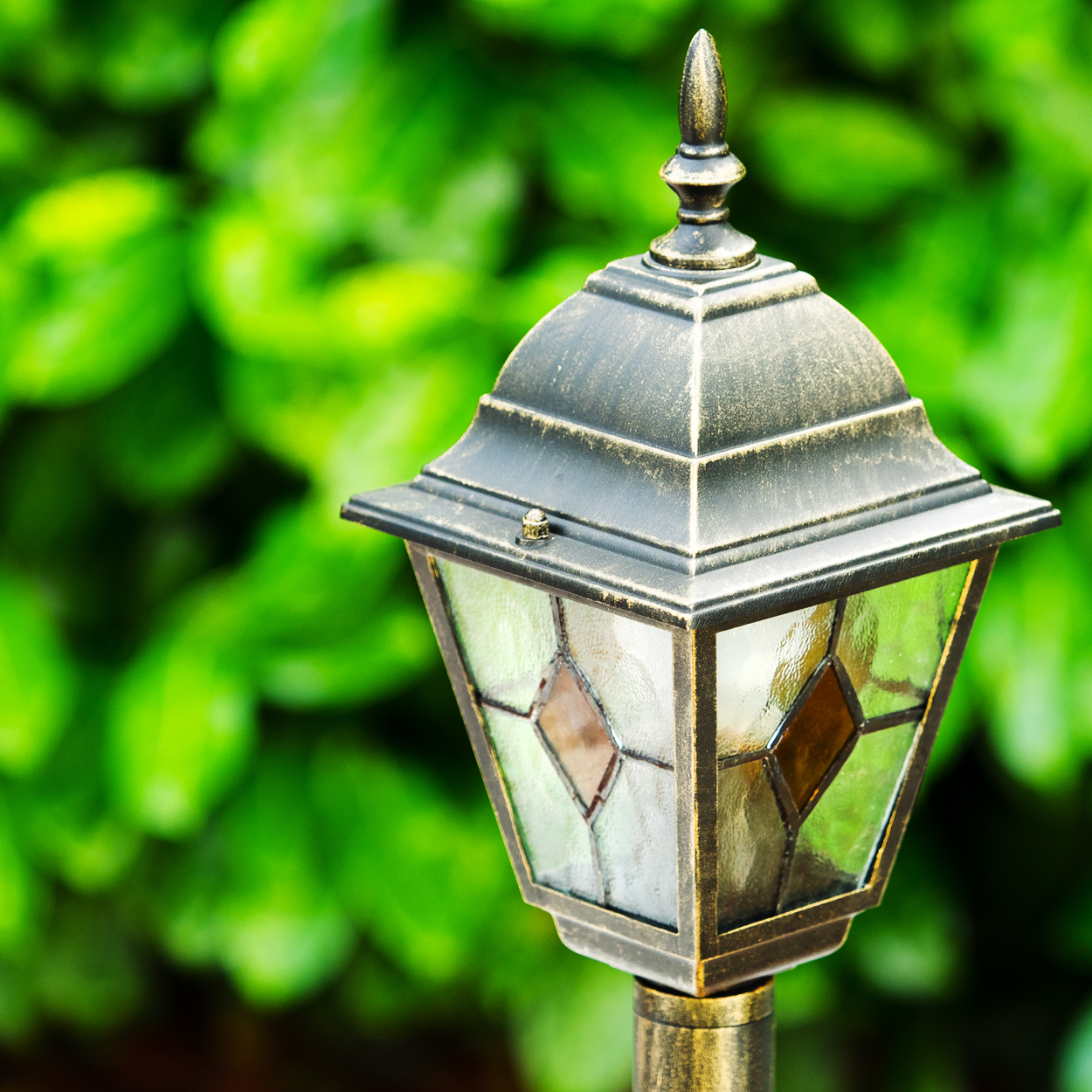 Eine verzierte Außenlampe mit Buntglasscheiben. Sie befindet sich vor einem Hintergrund aus verschwommenem grünem Laub.