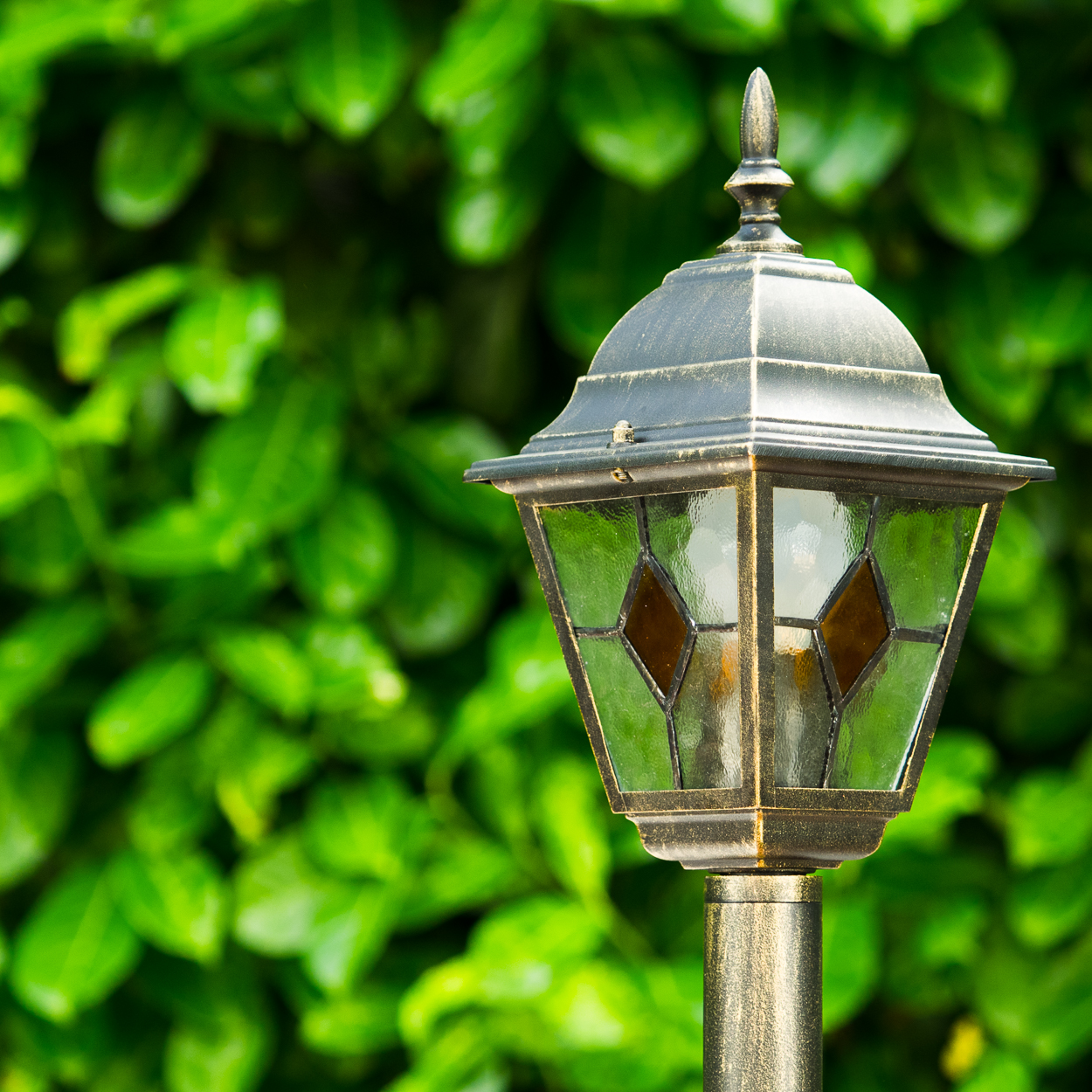 Eine dekorative Außenlampe mit Buntglasscheiben. Sie steht vor einem Hintergrund aus üppigen grünen Blättern.