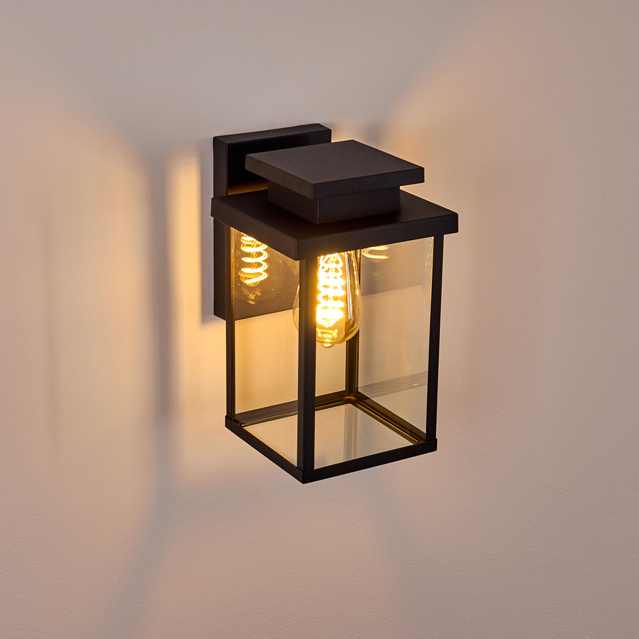 Eine schwarze Außenwandleuchte mit quadratischem Glasgehäuse und warmem Leuchtmittel. An einer beigen Wand montiert, wirft einen Schatten.
