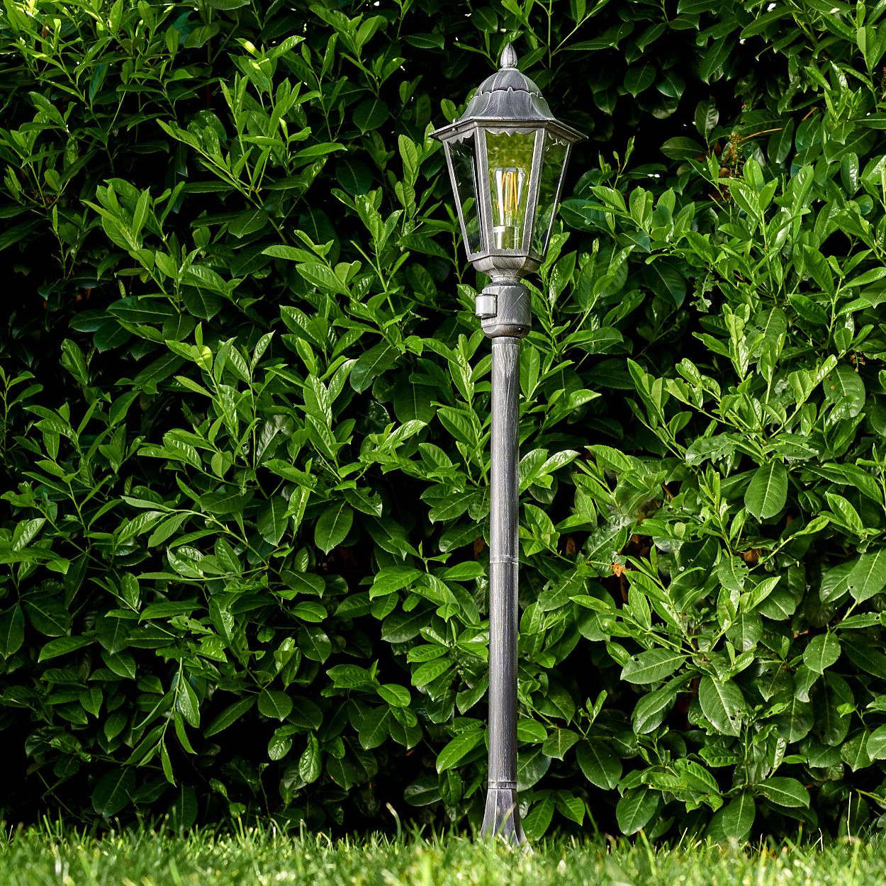 Eine graue Straßenlaterne steht vor einer grünen Hecke und grünem Gras. Die Lampe hat ein Glasgehäuse und das Licht ist an.