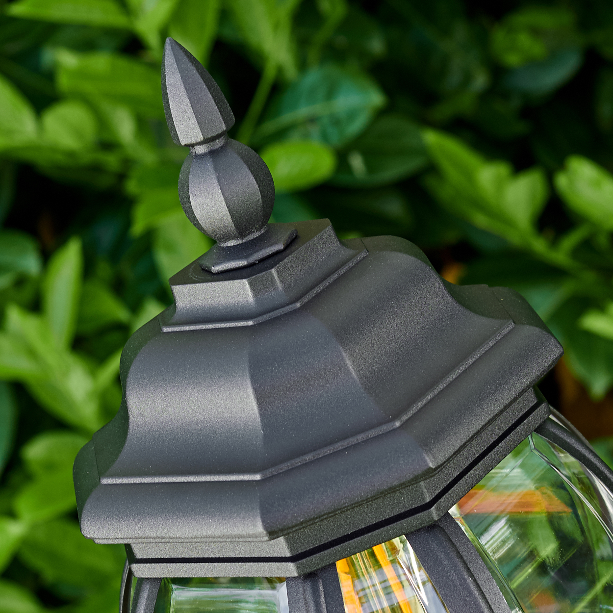 Nahaufnahme einer schwarzen Außenlampe mit verzierter Oberseite, vor einem Hintergrund aus grünen Blättern.