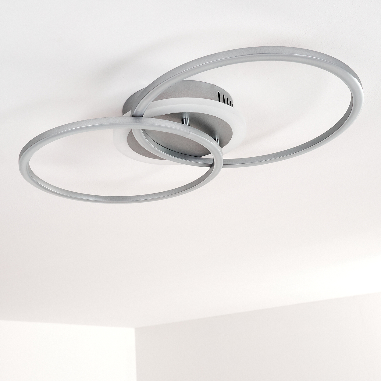 Eine moderne Deckenleuchte mit sich überlappenden silbernen Ringen und einer zentralen weißen Lichtquelle, vor weißem Hintergrund.