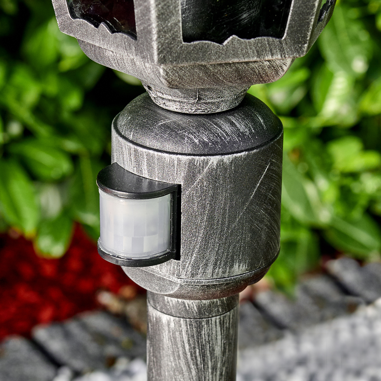 Nahaufnahme einer grauen und schwarzen Außenlaterne mit Sensor. Grünes Laub im Hintergrund, roter Mulch und Steine sind sichtbar.
