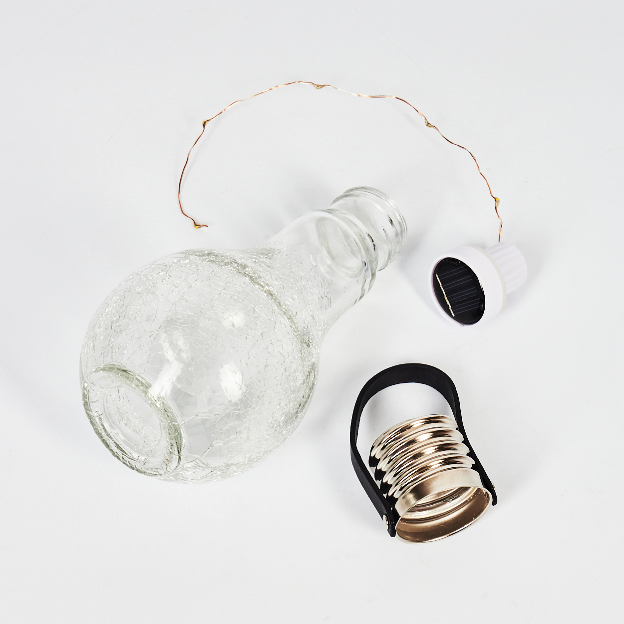 Eine zerlegte Glasflasche mit Solarverschluss, Metallring mit schwarzem Riemen und Lichterkette auf weißer Oberfläche.