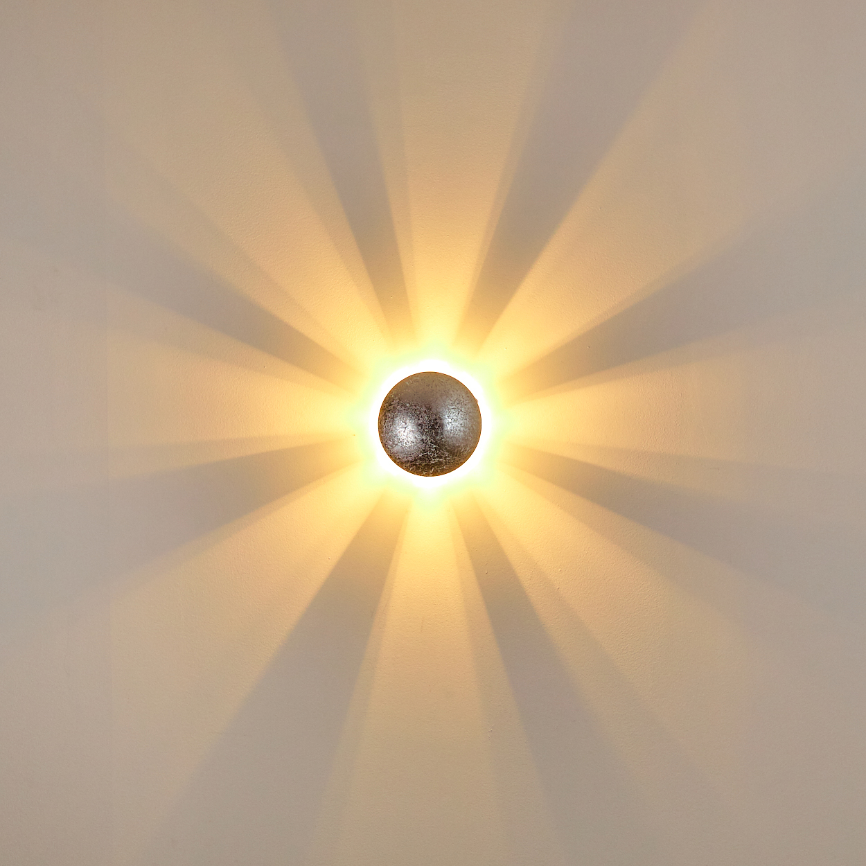 Eine runde, metallische Wandlampe strahlt Licht aus und wirft radiale Schatten auf eine hellfarbige Wand. Die Lichtquelle ist mittig.