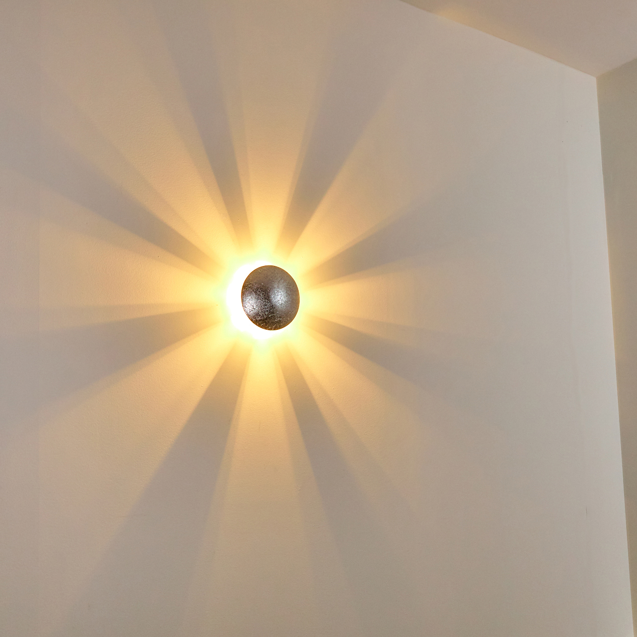 Eine runde, strukturierte silberne Wandleuchte strahlt Licht aus und wirft sonnenstrahlenartige Schatten auf die weiße Wand. Die Szene ist in Innenräumen.