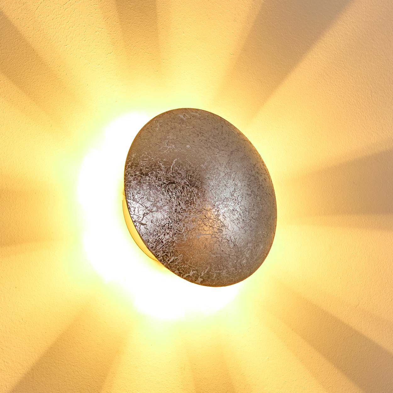 Eine silberne, kreisförmige Wandleuchte strahlt hellgelbes Licht aus. Das Licht verteilt sich auf der beigefarbenen Wand und erzeugt einen strahlenden Effekt.