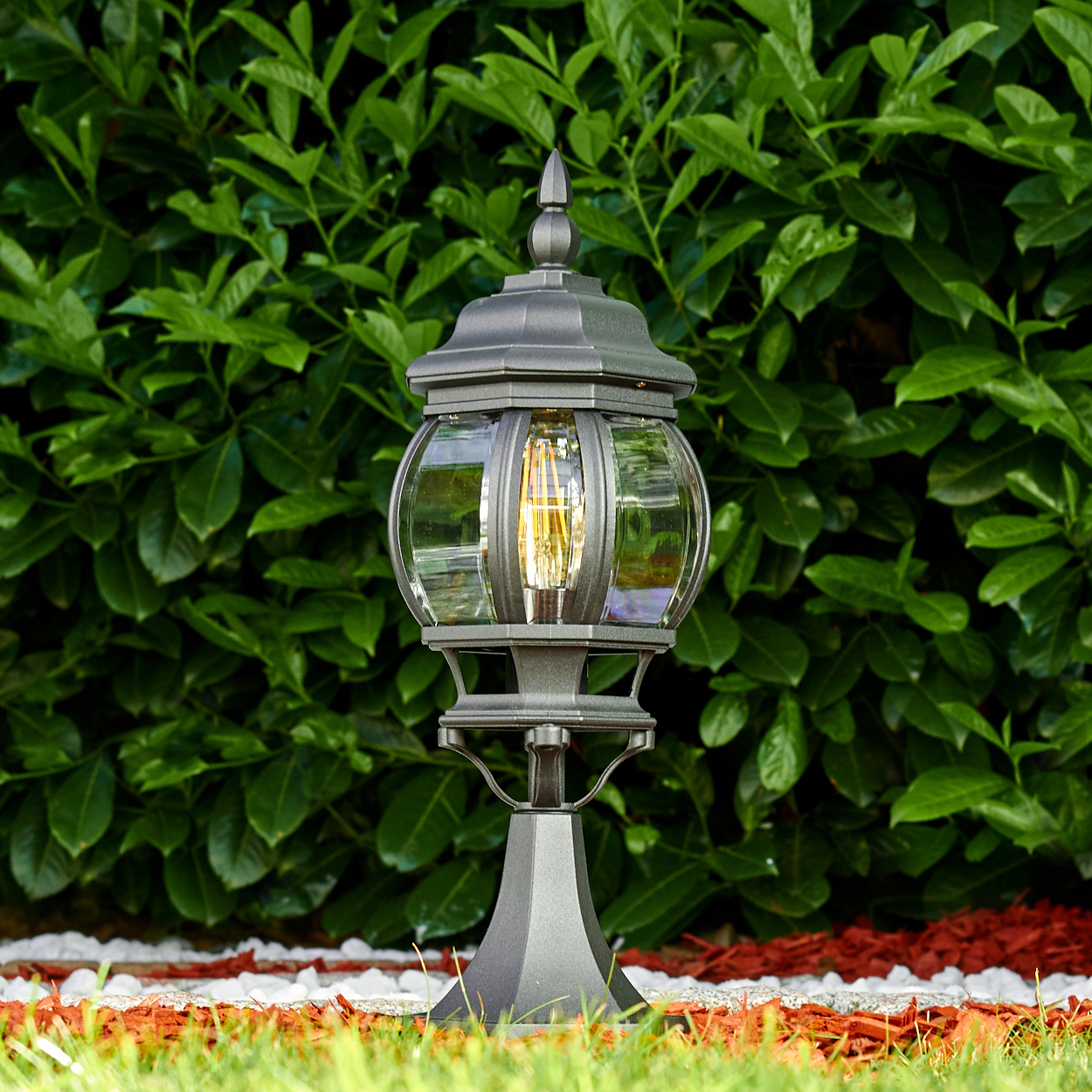 Eine dunkelgraue Außenlampe mit leuchtender Glühbirne steht auf Gras. Im Hintergrund grüne Büsche, am Sockel roter Mulch und weiße Steine.