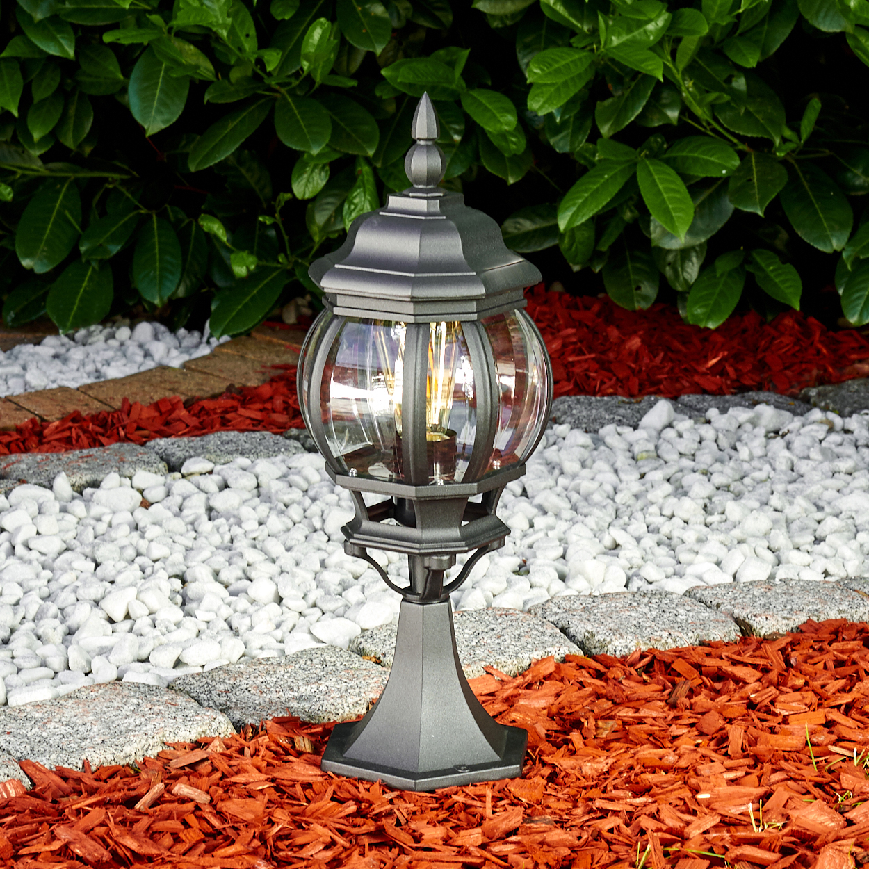 Eine schwarze Außenlampe steht in einem Garten mit roter Mulch, weißen Steinen und grünem Laub im Hintergrund. Die Lampe ist beleuchtet.
