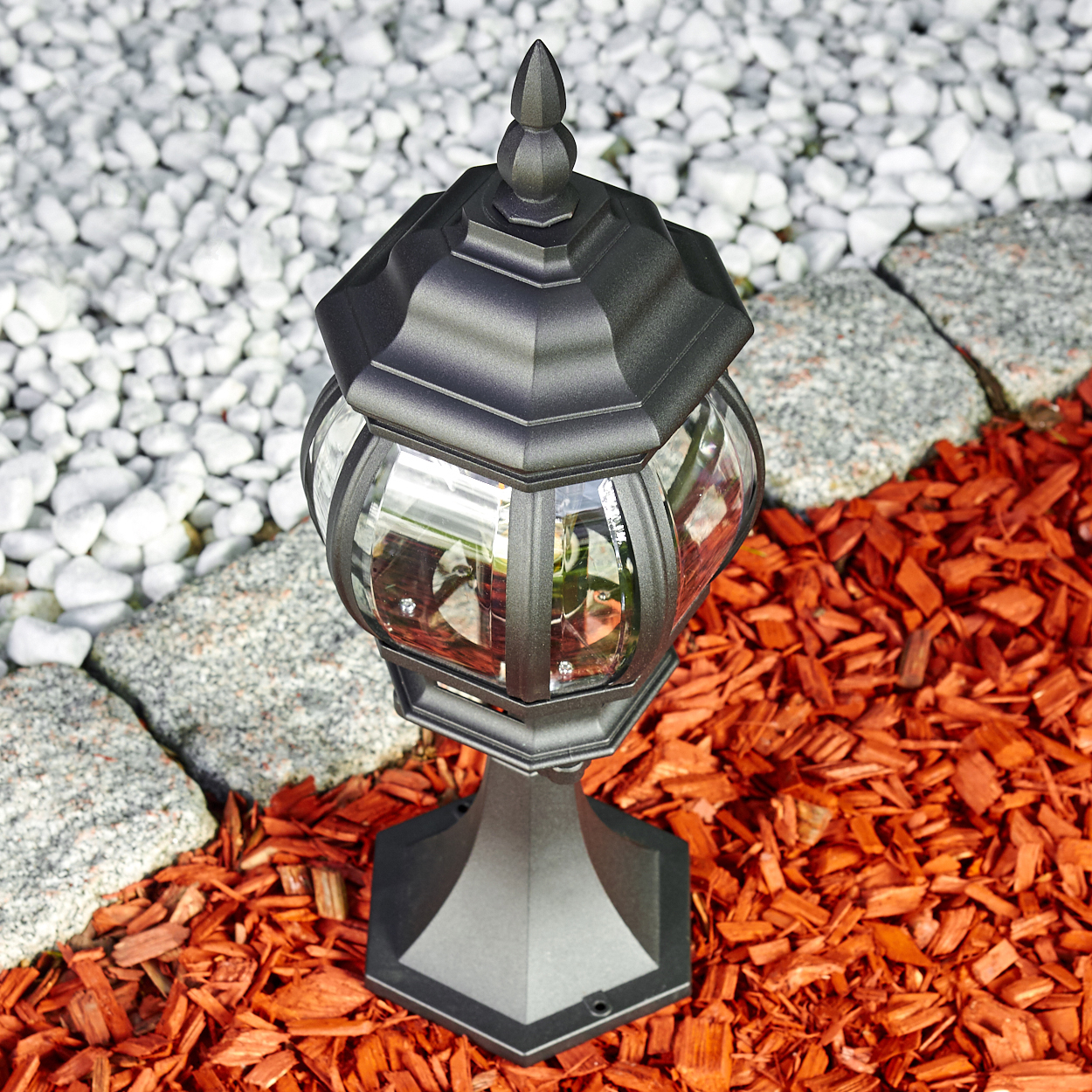 Eine schwarze Außenlampe mit Glasscheiben steht auf roter Mulch. Im Hintergrund befinden sich weiße Steine und graue Steine.