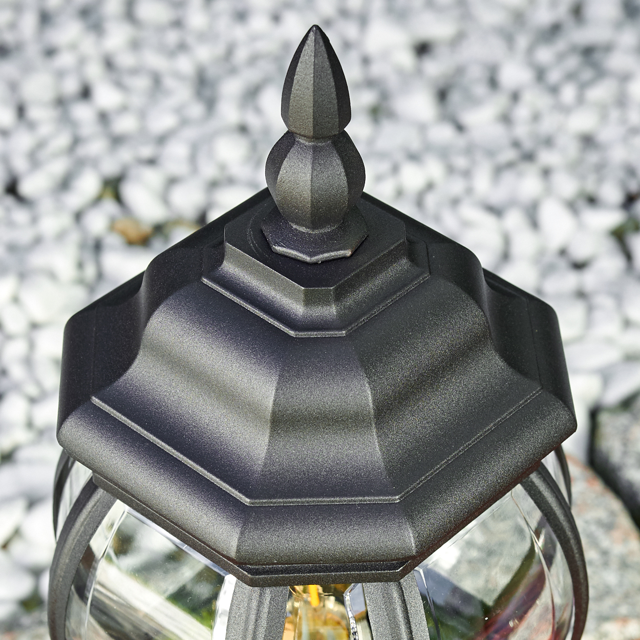 Nahaufnahme einer schwarzen Außenlampe. Die Lampe hat eine dekorative Oberseite und ein Glasgehäuse. Der Hintergrund besteht aus weißen Steinen.