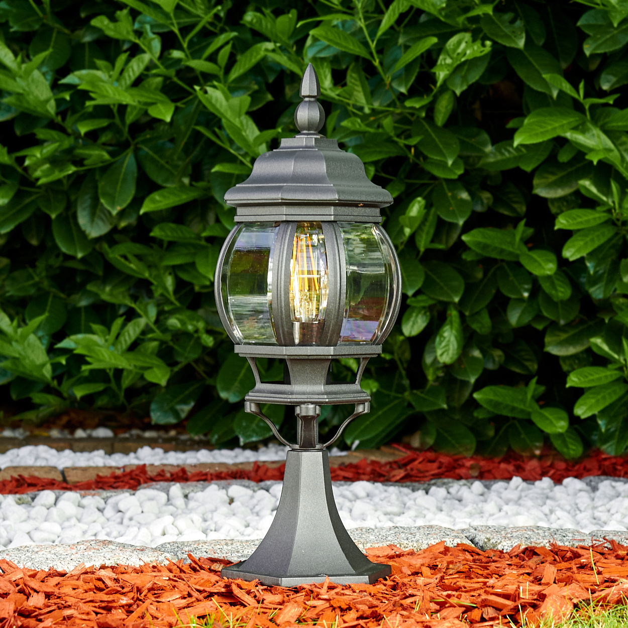 Eine schwarze Außenlampe mit einer Glühbirne, auf roter Mulch und weißen Steinen, grüne Büsche im Hintergrund.