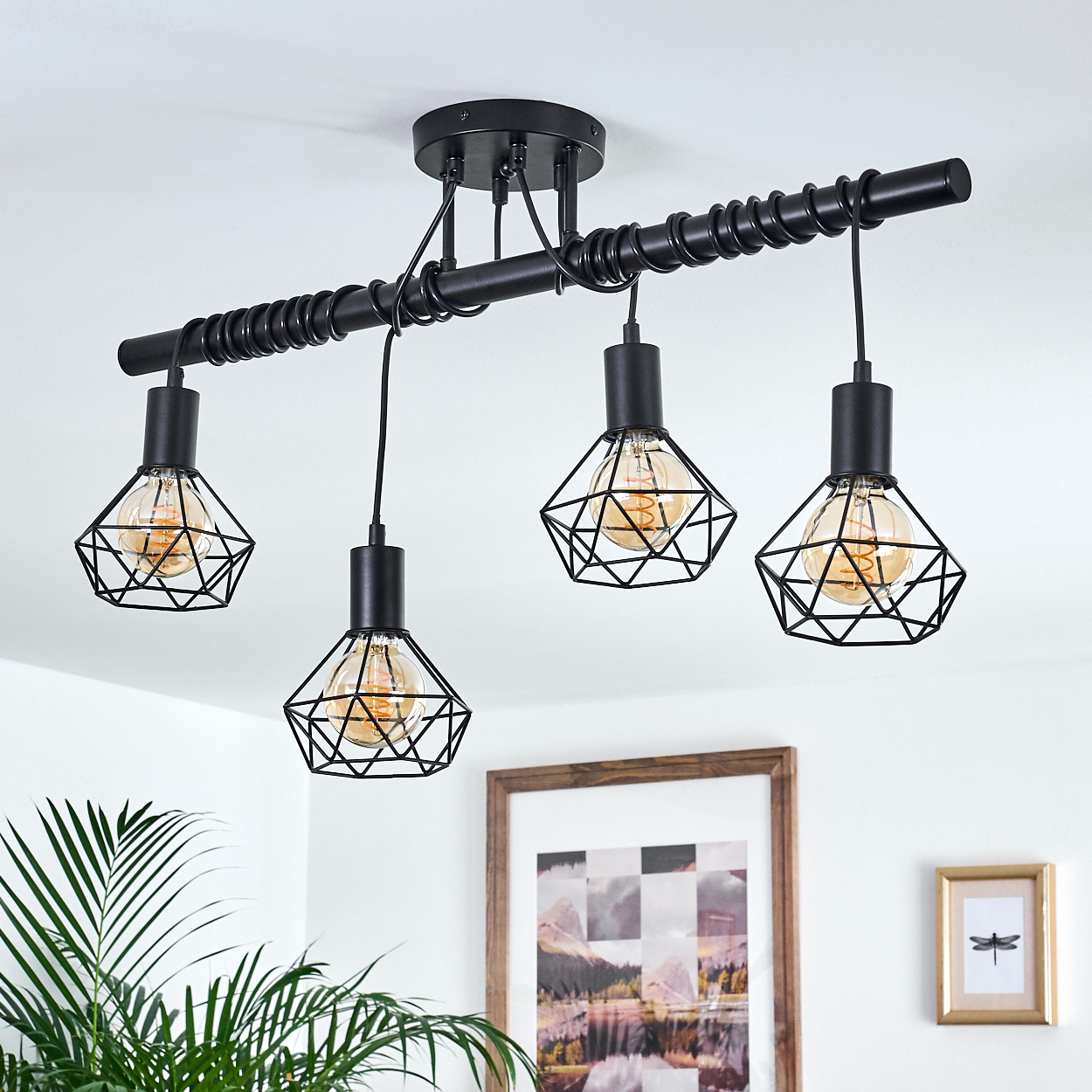 Schwarze, geometrische Pendelleuchten hängen von der Decke. Ein gerahmtes Bild und eine Pflanze befinden sich im Hintergrund. Die Lichter sind an.