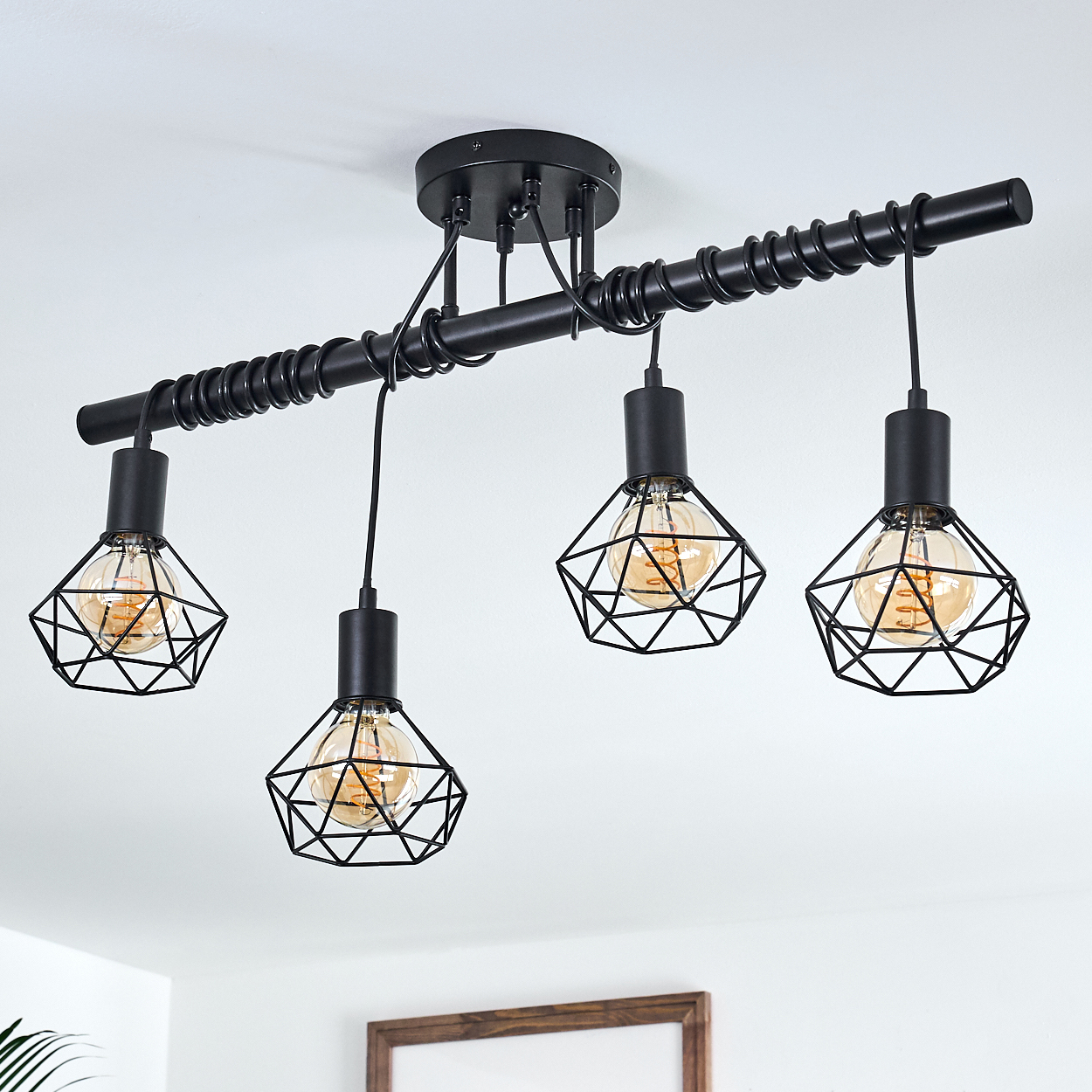 Schwarze geometrische Pendelleuchte mit vier Glühbirnen. Jede Glühbirne ist in einem Drahtkäfig eingeschlossen. An einer weißen Decke montiert.