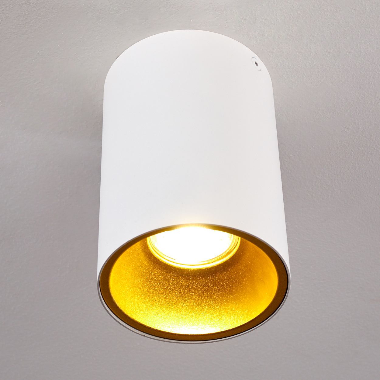 Eine weiße zylindrische Leuchte hängt an einer grauen Decke. Das Innere ist golden, und das Licht scheint.