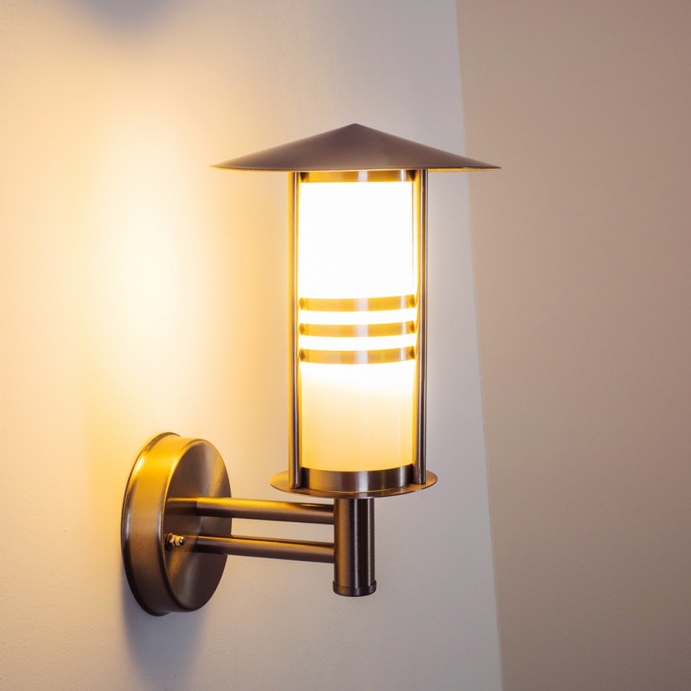 Eine Wandlampe mit zylindrischem Glasschirm und Metallrahmen. Die Lampe leuchtet und wirft ein warmes Licht an die Wand.