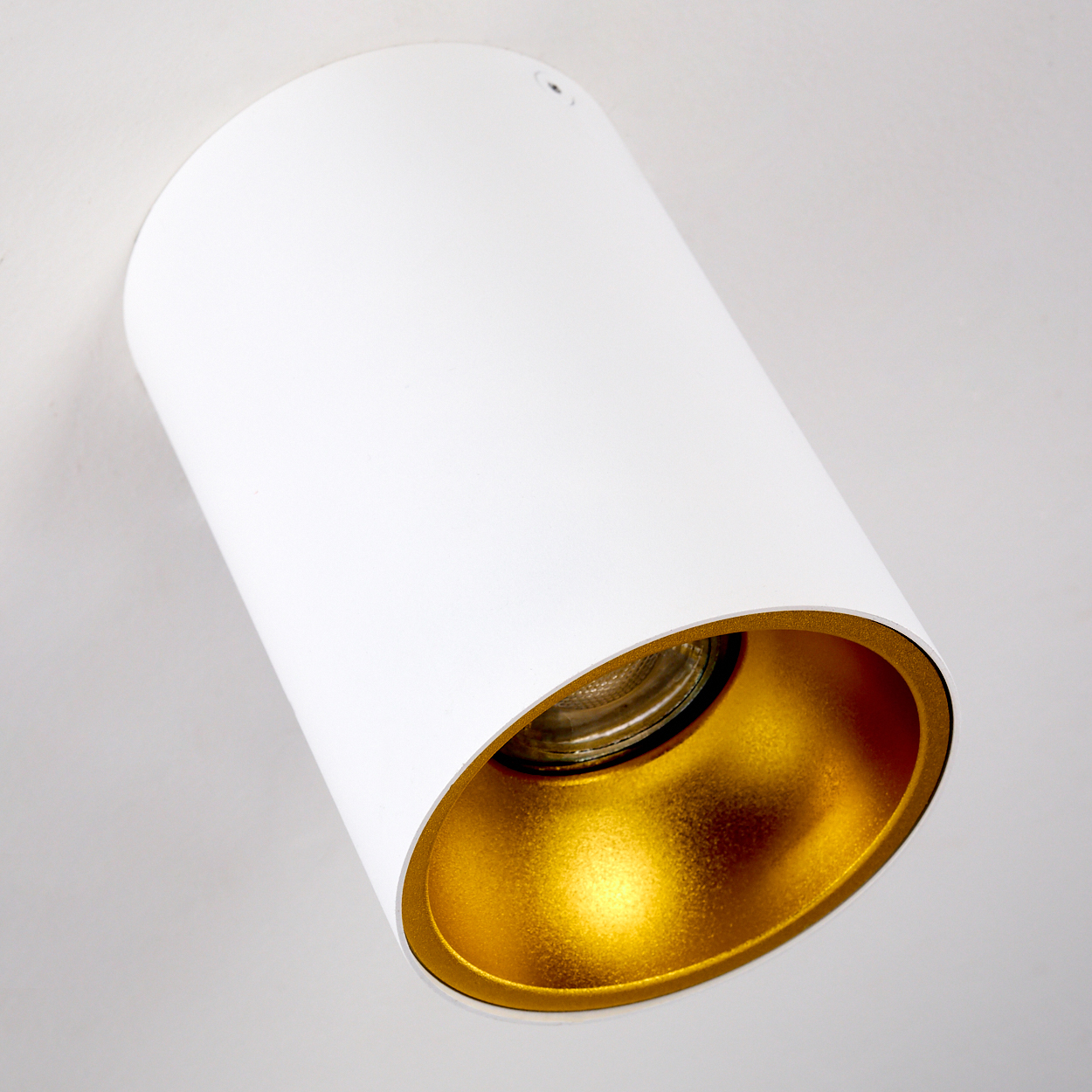 Eine weiße zylindrische Leuchte mit goldfarbenem Innenraum. Sie ist vor einem weißen Hintergrund abgewinkelt.