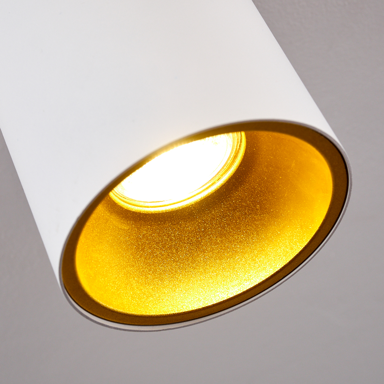 Nahaufnahme einer weißen zylindrischen Lampe mit goldenem Innenraum und hellem Licht. Der Hintergrund ist hellgrau.