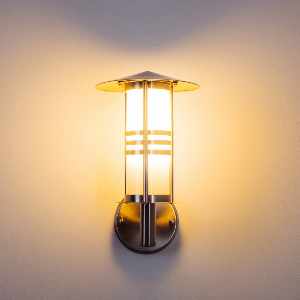 Eine wandmontierte, metallische Außenlampe leuchtet vor einer beigefarbenen Wand. Die Lampe hat ein zylindrisches Design mit horizontalen Streifen und einer konischen Spitze.