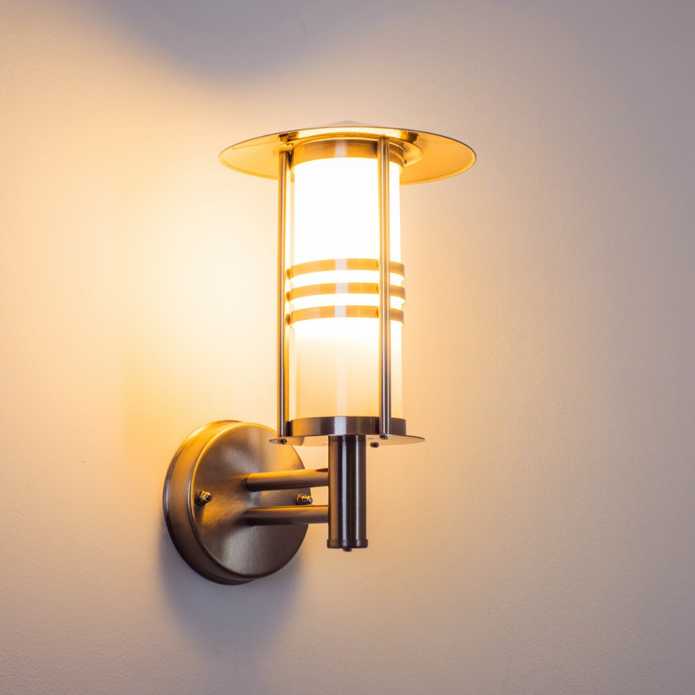 Eine Wandlampe mit zylindrischem Glasschirm, Metallrahmen und kreisförmiger Basis, die warmes Licht gegen eine helle Wand ausstrahlt.