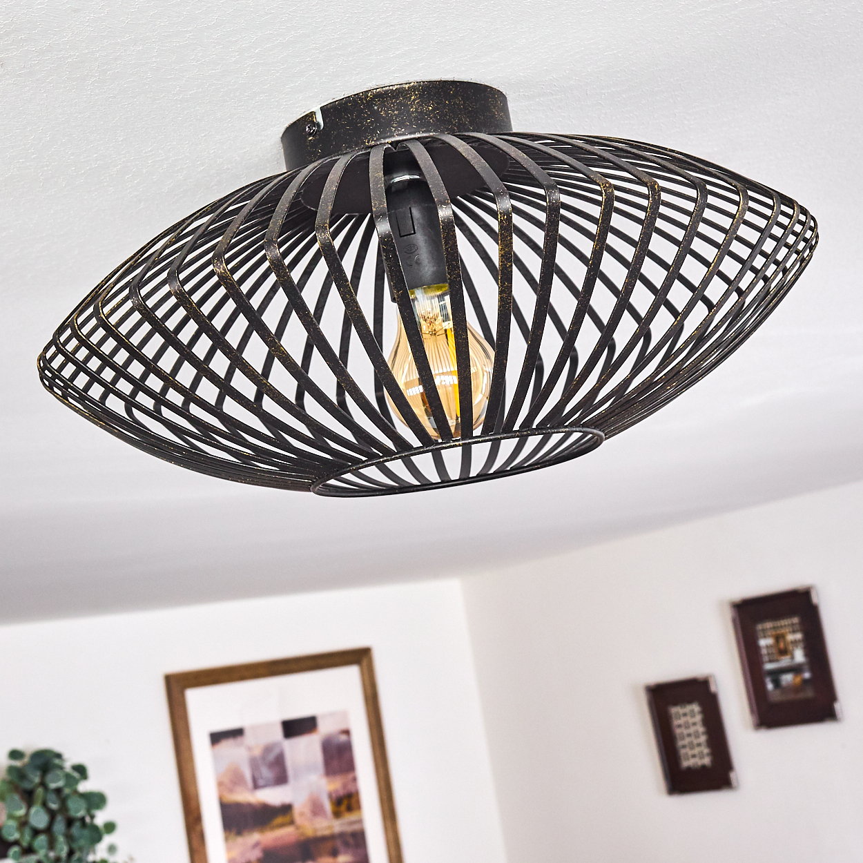 Schwarze Metallkäfig-Leuchte mit Glühbirne. Weiße Decke und Wand mit gerahmten Bildern.