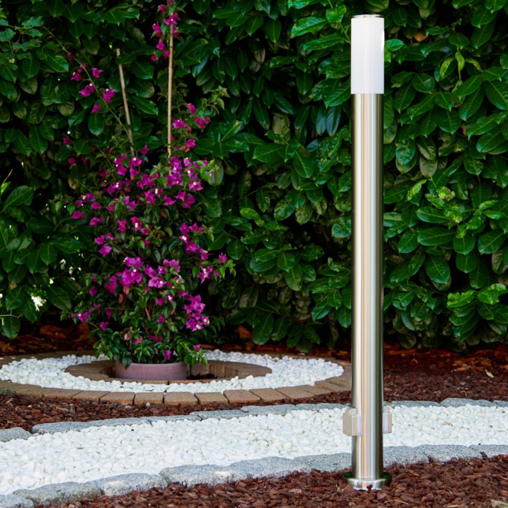 Eine Gartenszene mit einem silbernen Laternenpfahl. Links ist ein Busch mit lila Blüten. Weiße Steine bilden einen Weg.