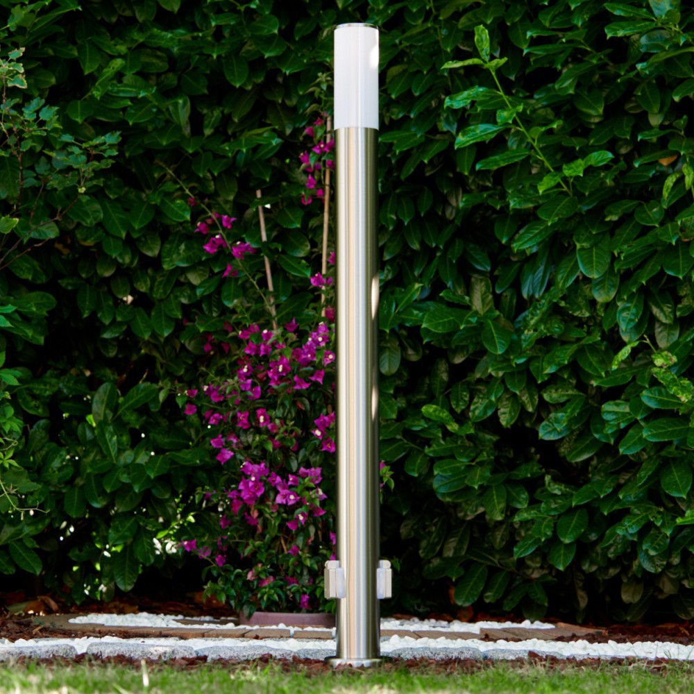 Eine Metallpolle steht in einem Garten. Dahinter befinden sich eine grüne Hecke und lila Blumen. Die Lampe hat einen weißen Aufsatz.