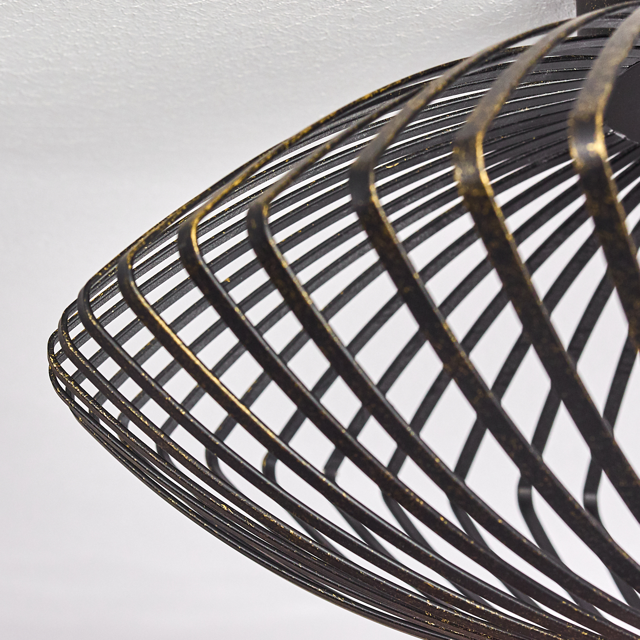 Ein schwarz-goldener Lampenschirm aus Metall mit Spiraldesign hängt vor weißem Hintergrund. Die Nahaufnahme zeigt die Streben.