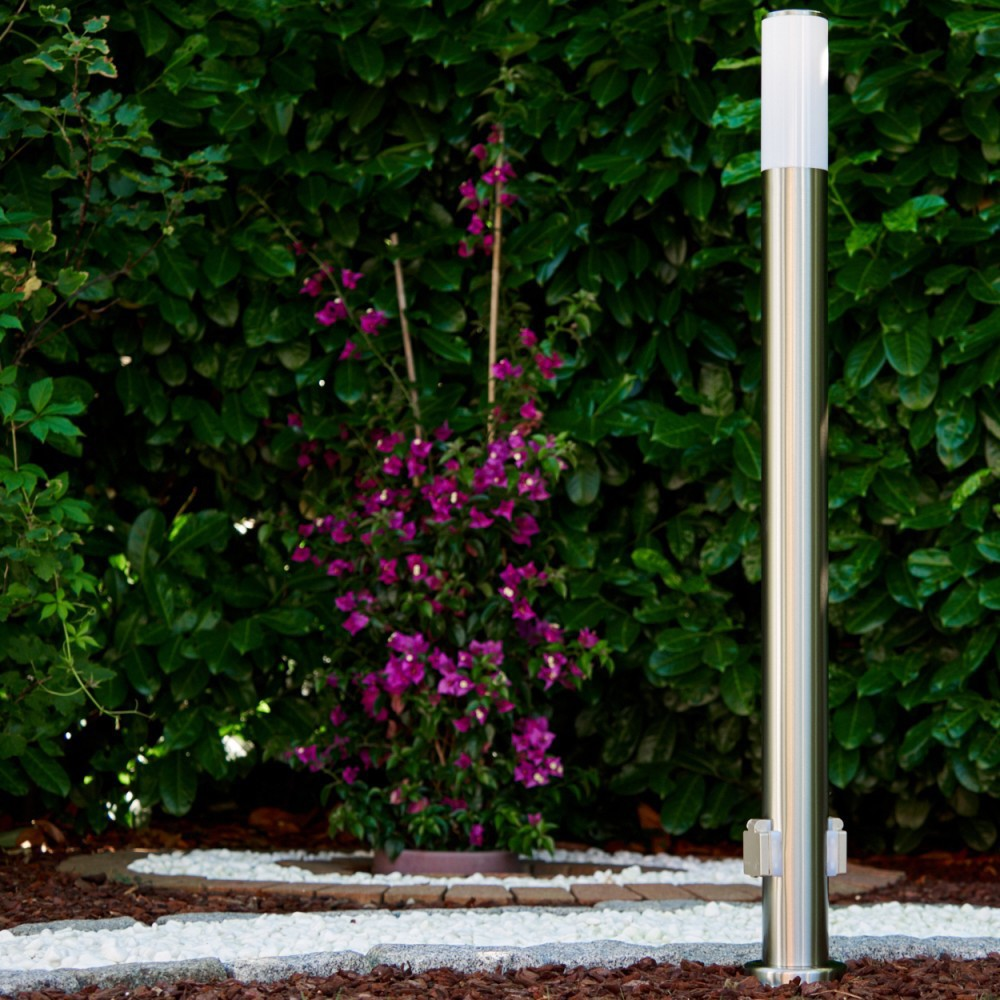 Ein Edelstahl-Lichtmast steht vor einer grünen Hecke. Links ist ein blühender Strauch mit lila Blüten.