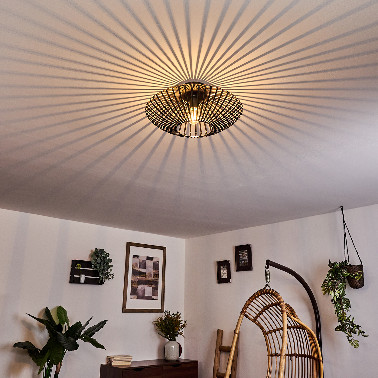 Eine Deckenleuchte wirft Schatten. Ein Schaukelstuhl aus Rattan, Pflanzen und gerahmte Bilder dekorieren den Raum.