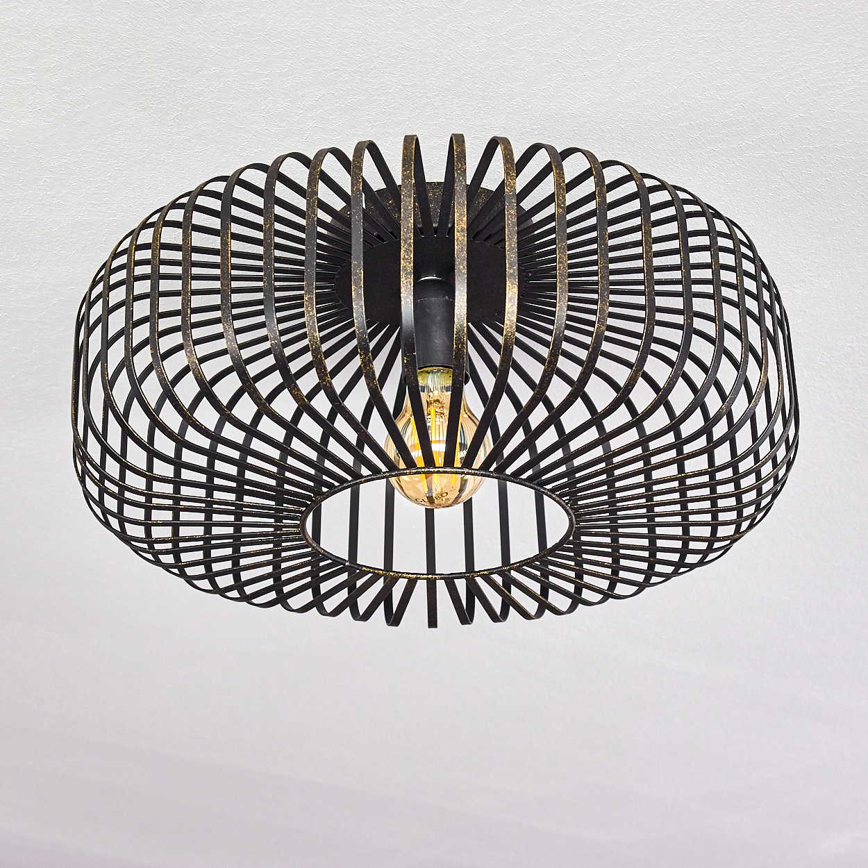 Eine schwarze Deckenlampe mit Drahtrahmen und einer leuchtenden Glühbirne. Die Lampe hat eine ovale Form mit geometrischem Design, vor weißer Decke.