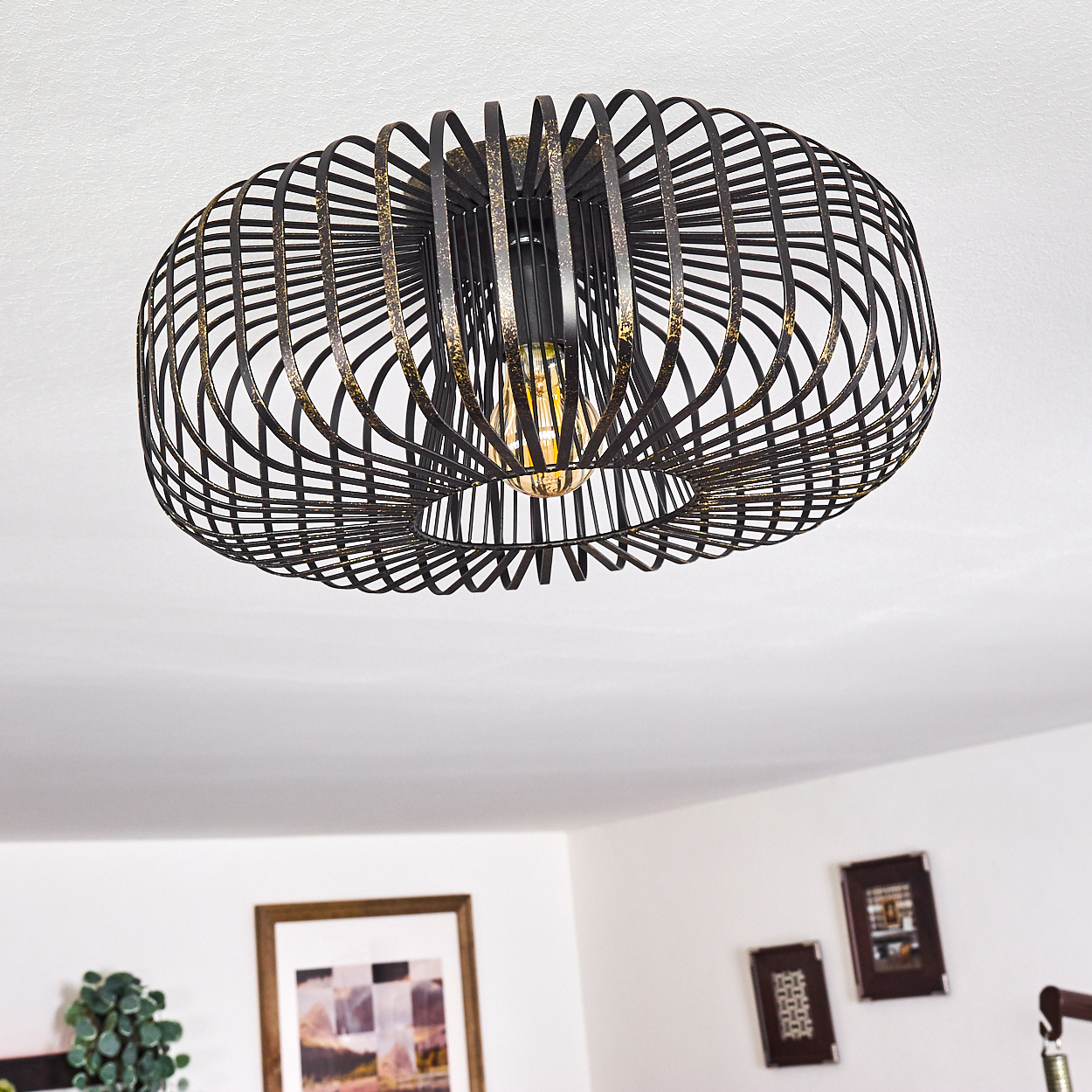 Moderne, schwarz-goldene Deckenleuchte mit käfigartigem Design. Das Licht ist an und beleuchtet den Raum. Andere Rahmen sind sichtbar.