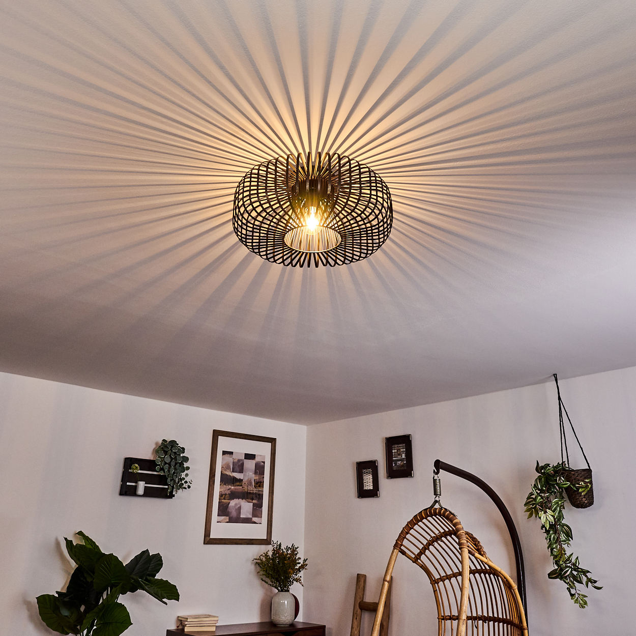 Eine schwarze, geometrische Deckenlampe wirft radiale Schatten. Darunter ist ein Raum mit Pflanzen, Kunst und einem Hängestuhl zu sehen.