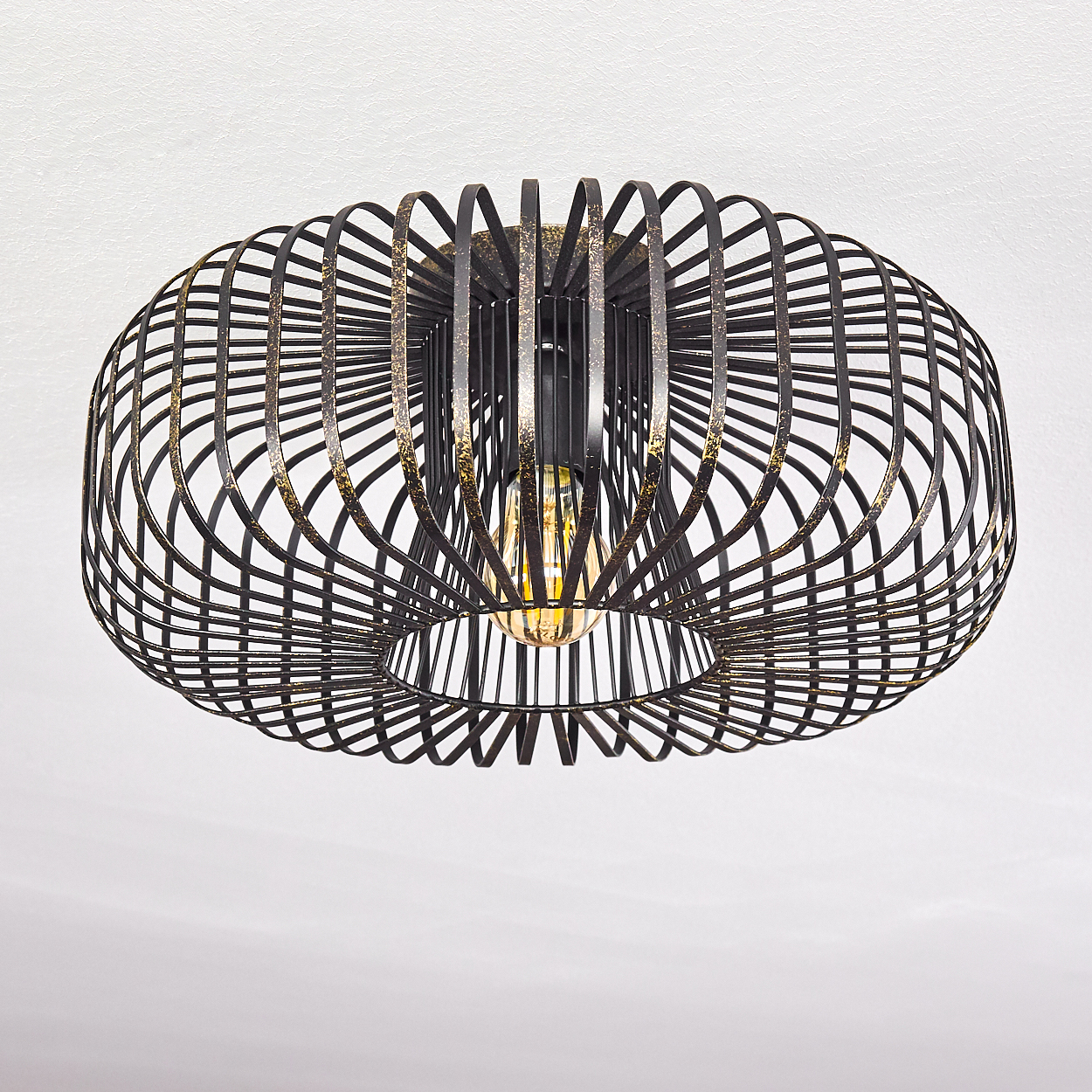 Eine verzierte, dunkle Metallleuchte. Die Leuchte hat ein käfigartiges Design mit einer sichtbaren Glühbirne im Inneren. Sie hängt an einer weißen Decke.
