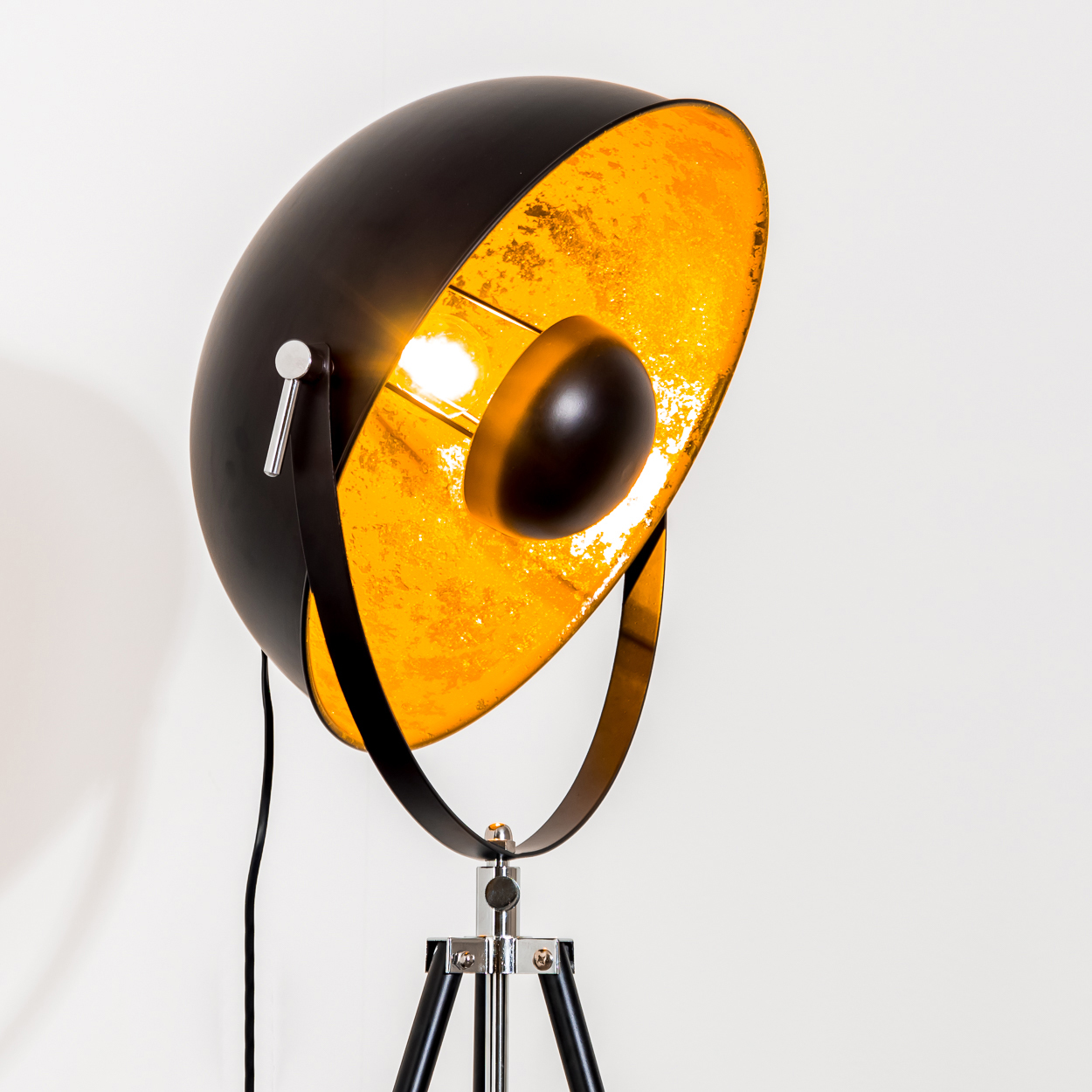Eine schwarz-goldene Stehlampe auf einem Stativsockel vor weißem Hintergrund. Die Lampe ist eingeschaltet.