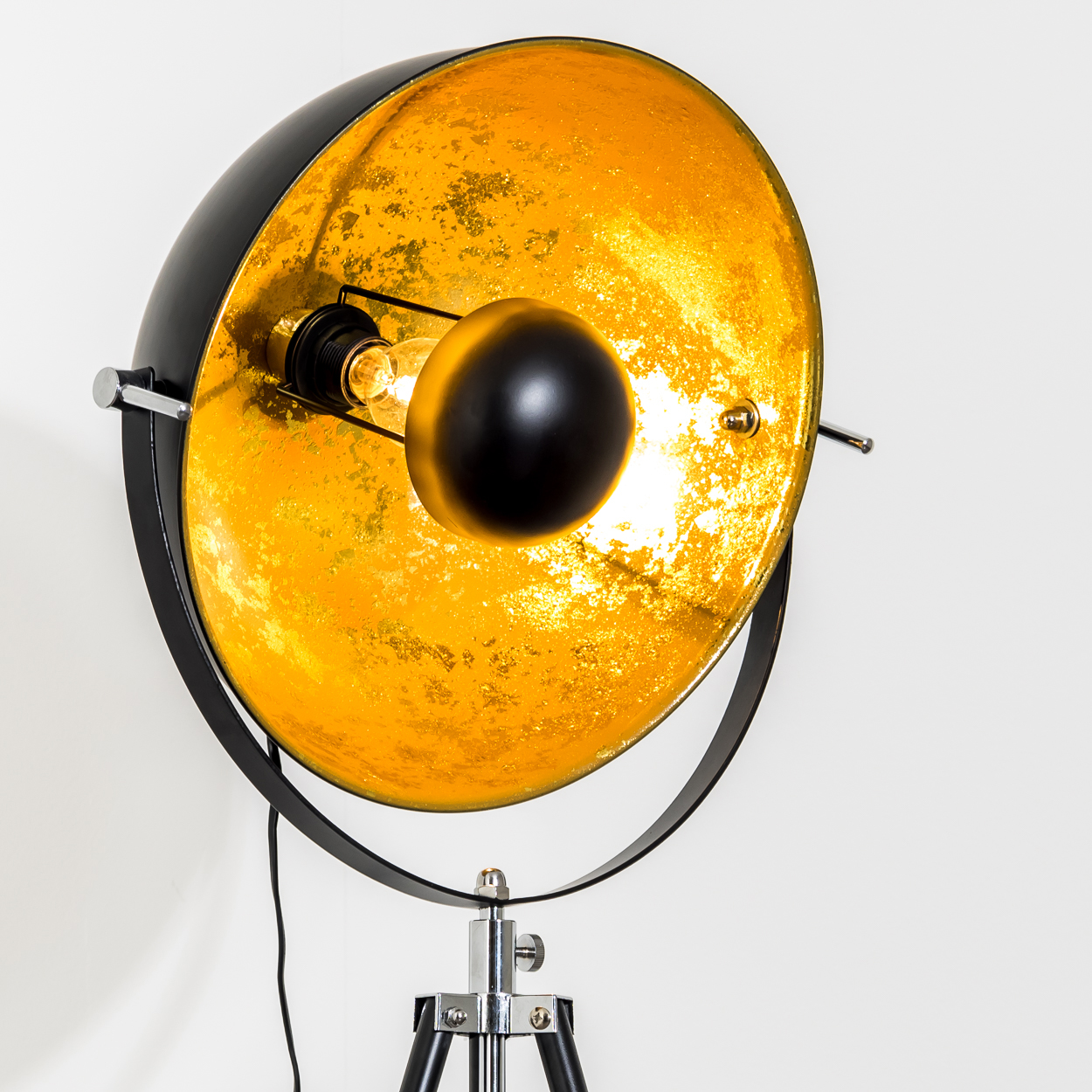 Eine schwarz-goldene Stehlampe mit rundem Schirm und Stativfuß. Die Glühbirne leuchtet und der Hintergrund ist weiß.