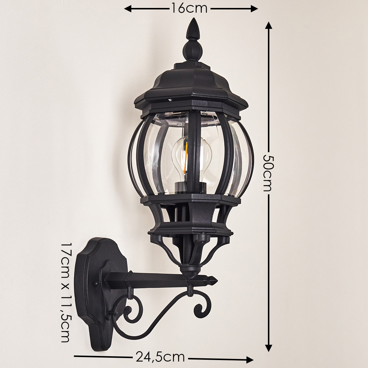 Eine schwarze, an der Wand montierte Außenlampe. Sie hat einen Glaskolben, eine verzierte Halterung und befindet sich an einer cremefarbenen Wand.