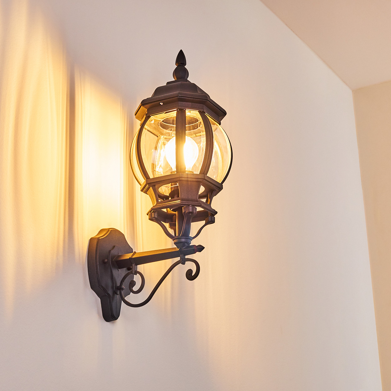 Eine verzierte, schwarze Wandlampe mit Glaskugel leuchtet und wirft einen warmen Schein auf die cremefarbene Wand. Die Lampe ist in der Nähe einer Ecke montiert.