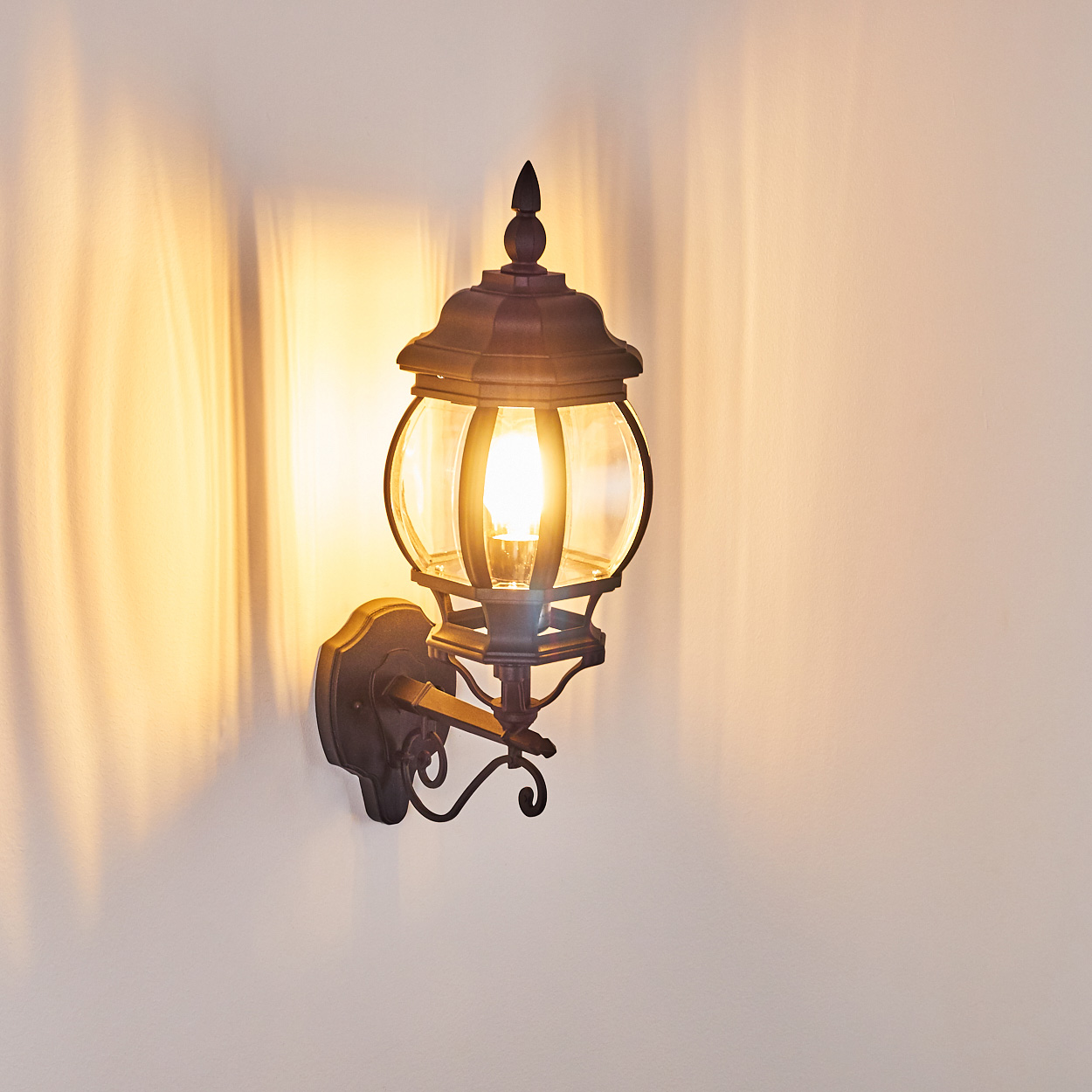 Eine verzierte Außenwandleuchte mit leuchtender Glühbirne ist an einer weißen Wand befestigt und wirft weiche Schatten. Die Lampe ist dunkelbraun.