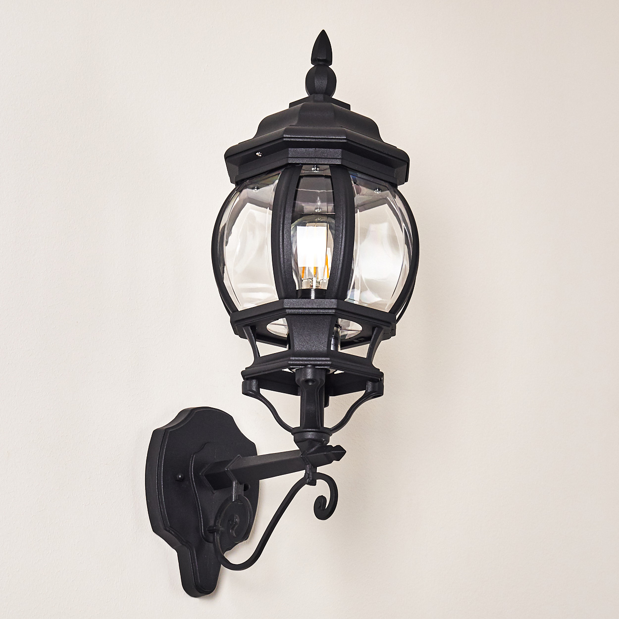 Eine schwarze, an der Wand montierte Außenlampe mit Glaseinfassung. Die Lampe befindet sich an einer hellbeige Wand. Im Glas ist eine Glühbirne sichtbar.