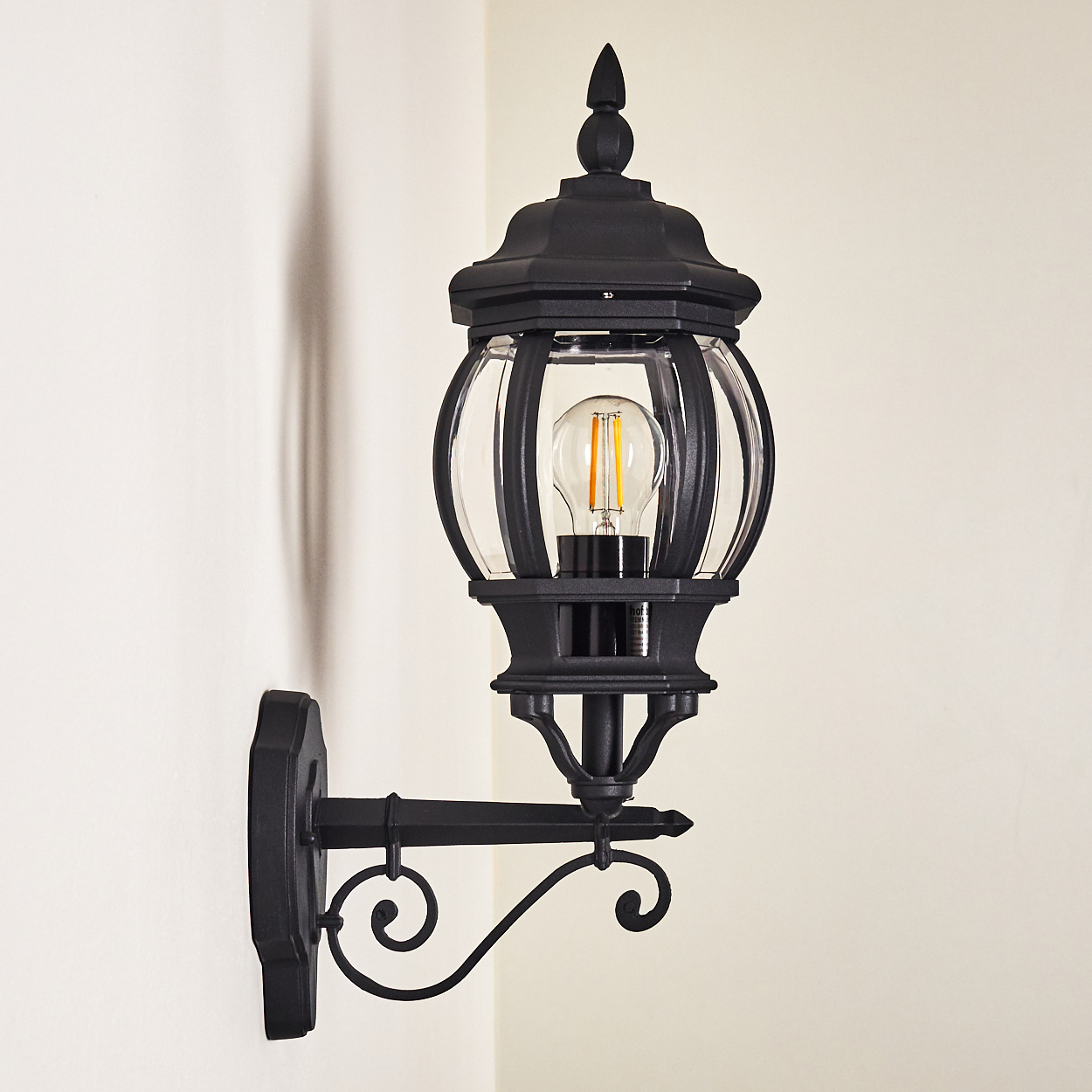 Eine schwarze Außenlampe ist an einer weißen Wand montiert. Die Lampe hat eine Glaskugel und einen dekorativen Arm.