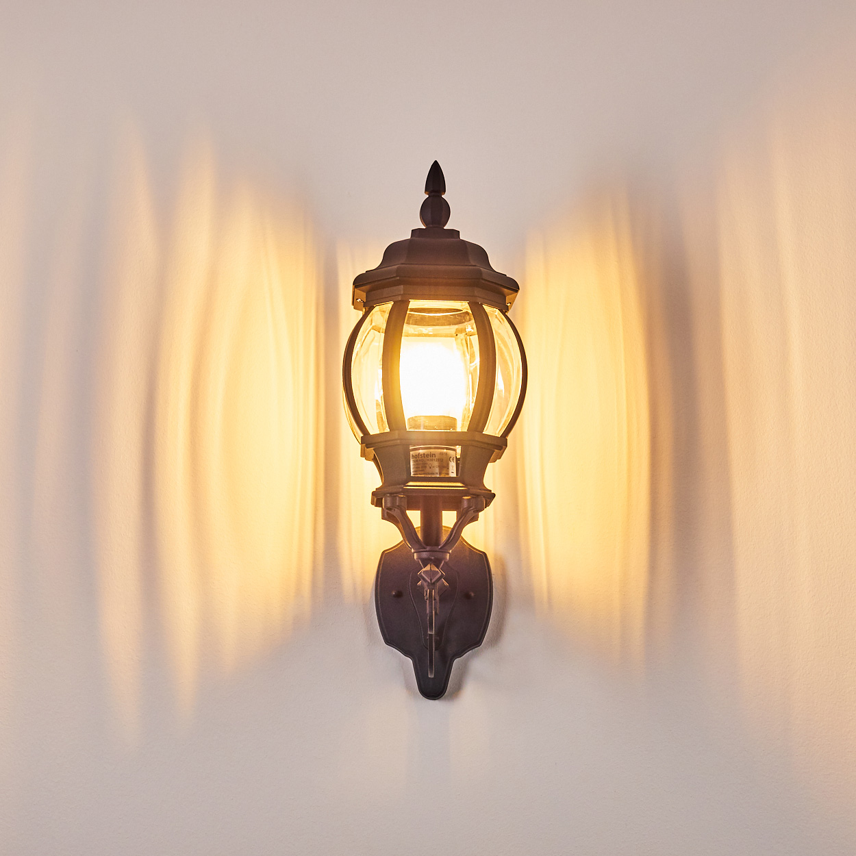 Eine verzierte schwarze Außenlampe ist an einer weißen Wand befestigt. Die Lampe ist beleuchtet und wirft warmes Licht und Schatten.