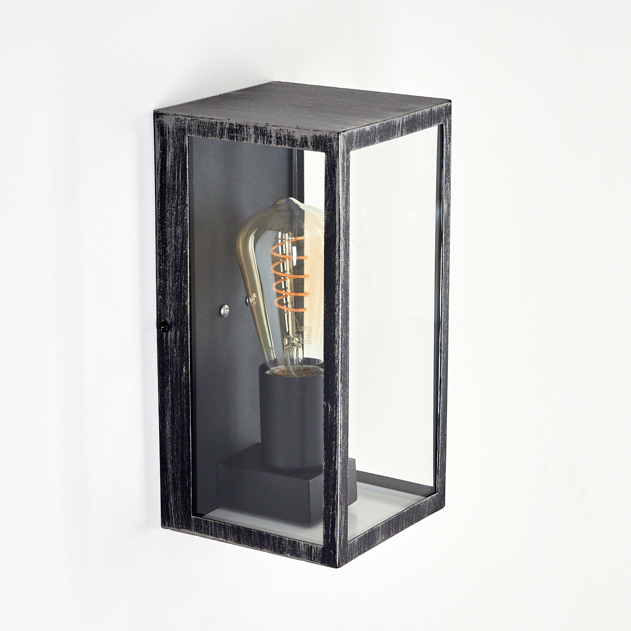 Eine schwarz-silberne Außenwandleuchte mit Glasgehäuse. Im Inneren befindet sich eine Edison-Glühbirne. Die Lampe ist an einer weißen Wand montiert.